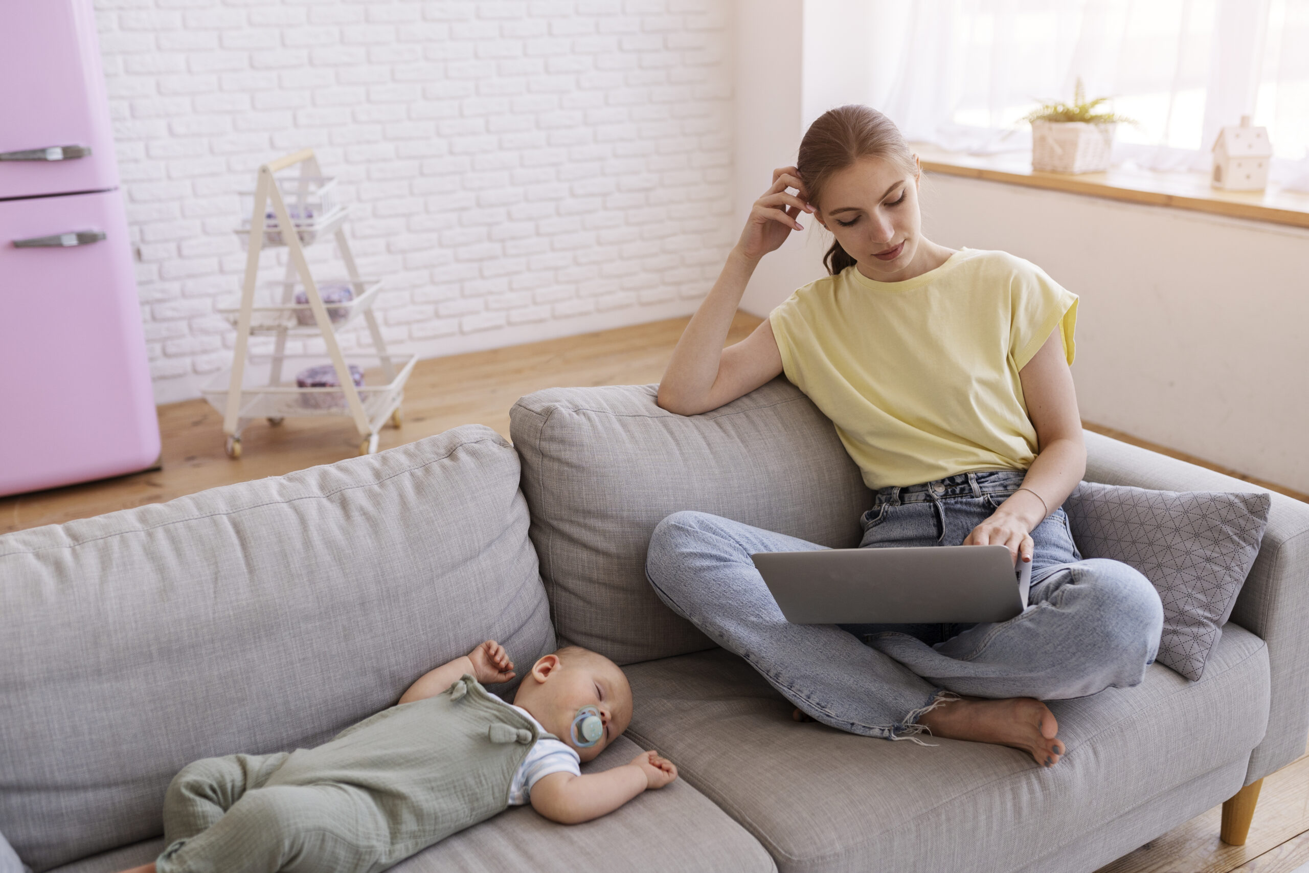Young mother working on a laptop while sitting on a couch next to her sleeping baby, balancing work and motherhood at home